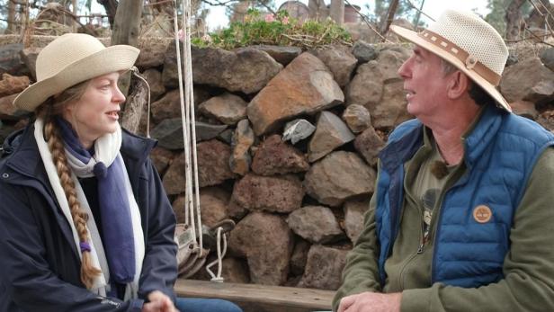 Ein Mann und eine Frau sitzen vor einer Steinmauer und unterhalten sich.