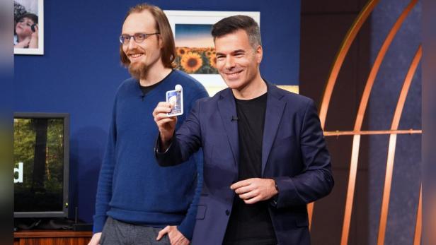 Zwei Männer stehen vor einer blauen Wand, einer hält eine Karte der „Höhle der Löwen“ hoch.