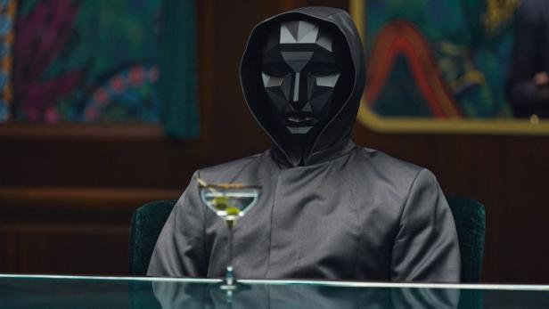 Eine Person mit einer schwarzen Maske und einem grauen Kapuzenpullover sitzt an einem Tisch mit einem Martini.