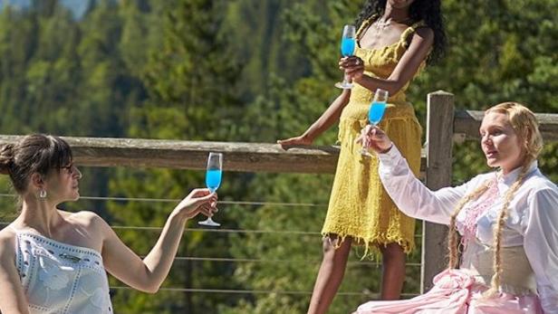 Drei Frauen stoßen mit blauen Getränken vor einer Bergkulisse an.