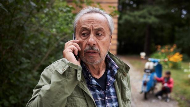Ein besorgter Mann telefoniert im Freien; im Hintergrund ein Kind und ein blauer Roller.