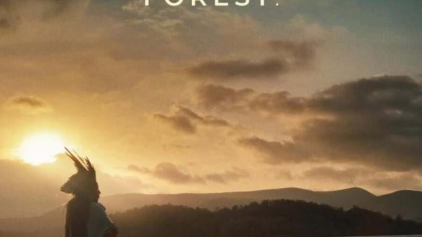 Ein Mann mit Kopfschmuck steht am Ufer eines Sees bei Sonnenuntergang, im Vordergrund der Filmtitel „Yanuni“.