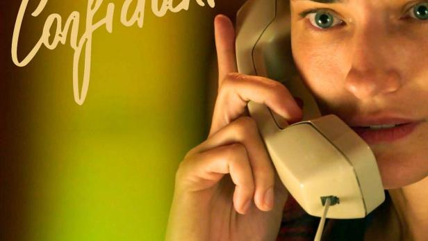 Eine Frau telefoniert mit einem alten Telefonhörer vor dem Schriftzug „Confidante“.