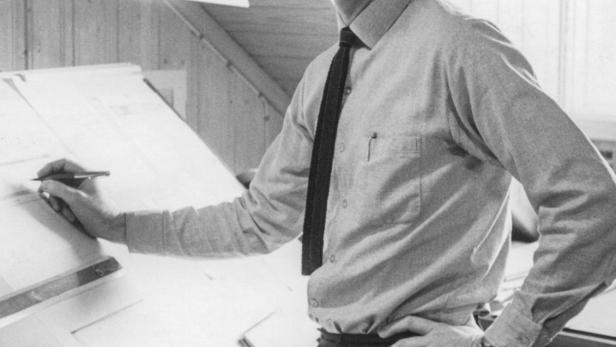 Ein Mann steht an einem Schreibtisch und zeichnet mit einem Stift auf ein Blatt Papier.