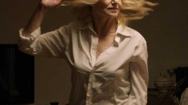 Eine Frau mit blonden Haaren trägt ein weißes Hemd und gestikuliert in einem dunklen Raum.