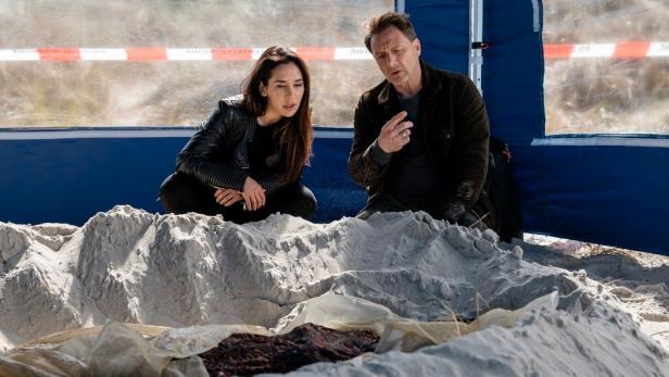 Zwei Personen untersuchen einen Leichnam im Sand, möglicherweise am Tatort eines Nordseekrimis.