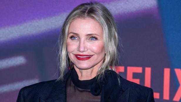Ein Porträt von Cameron Diaz mit rotem Lippenstift vor einem violetten Hintergrund.