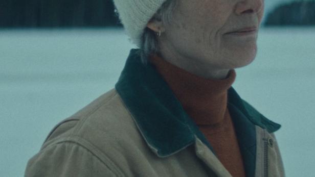 Eine ältere Frau mit Mütze und Jacke steht in einer verschneiten Landschaft.