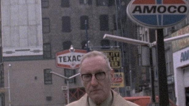 Ein älterer Mann mit Brille und Mantel steht vor einer Tankstelle in der Stadt.
