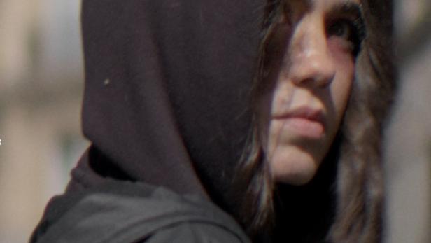 Eine junge Frau mit Kapuze und einem blauen Fleck unter dem Auge blickt in die Ferne.