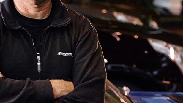 Ein Mann mit Brille steht vor einem blauen Oldtimer mit geöffneter Motorhaube.