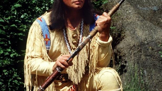 Ein Schauspieler als Winnetou, mit Gewehr, vor einem Felsen und Grünzeug.