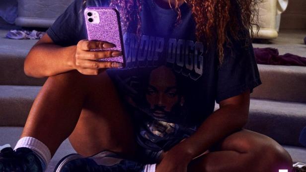 Eine junge Frau sitzt mit einem Glitzer-Handy in ihrem unordentlichen Zimmer.