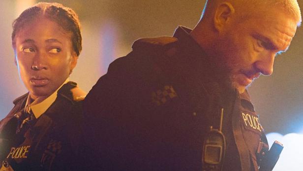 Ein Mann und eine Frau in Polizeiuniformen stehen Rücken an Rücken vor einem unscharfen Hintergrund.