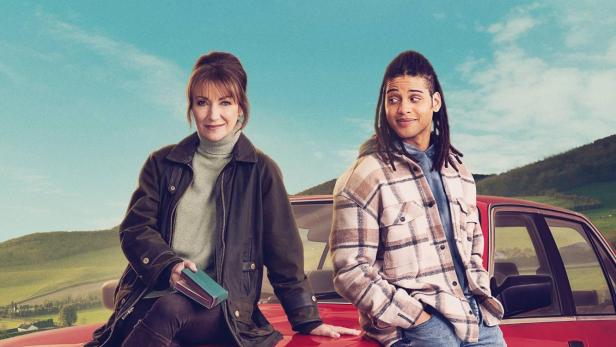 Die Schauspieler Jane Seymour und Rohan Nedd stehen vor einem roten Auto für „Harry Wild“.