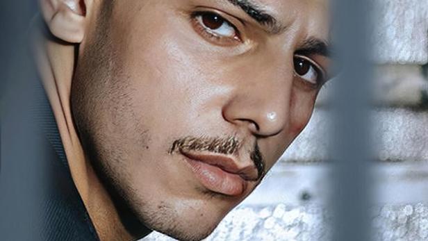 Ein Mann mit Schnurrbart blickt hinter Gittern hervor; im Vordergrund steht das Wort „ASBEST“.