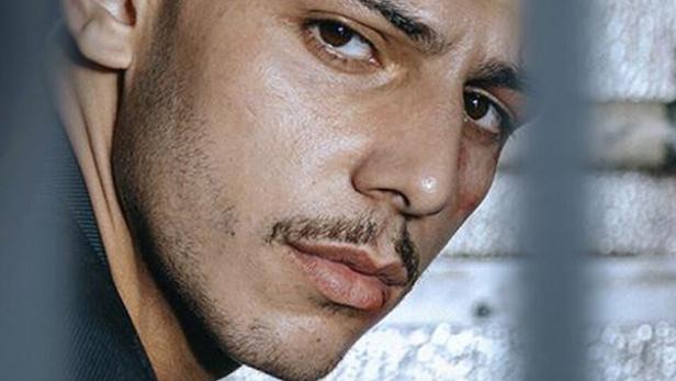 Ein junger Mann blickt hinter Gittern hervor; im Vordergrund steht das Wort „ASBEST“.