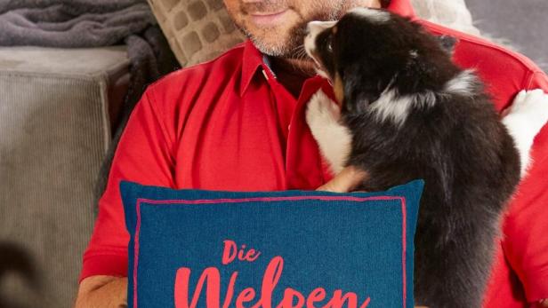 Martin Rütter hält einen Welpen vor einem Kissen mit der Aufschrift „Die Welpen kommen“.
