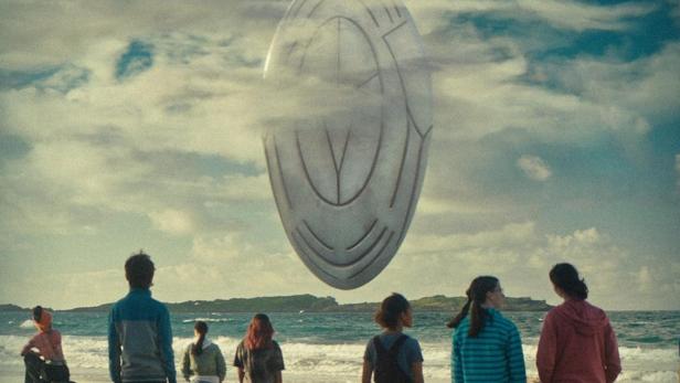 Eine Gruppe von Menschen am Strand blickt auf ein schwebendes, silbernes Objekt.