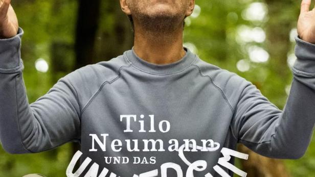 Tilo Neumann sitzt im Wald und blickt mit erhobenen Händen nach oben.
