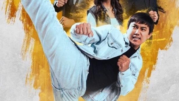 Ein Mann in Jeansjacke führt einen hohen Tritt vor dem Hintergrund weiterer Personen aus.