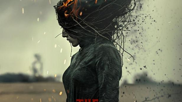 Eine Frau steht mit brennenden Ästen auf dem Kopf vor einem trüben Himmel.