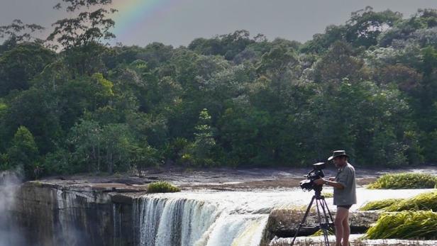 Ein Kameramann filmt einen Wasserfall vor einer tropischen Landschaft unter einem Regenbogen.