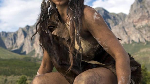 Eine junge Frau in Fellkleidung kauert vor einer Bergkulisse.