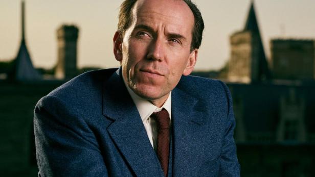 Ein Mann im Anzug und Handschuhen, vermutlich Professor T, vor einer Stadtkulisse.