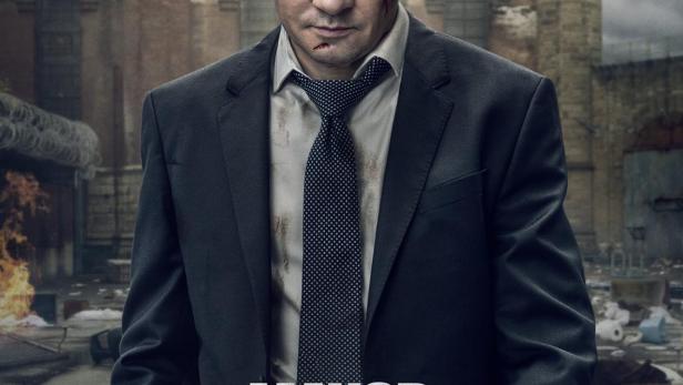 Ein verletzter Mann im Anzug steht vor einem Gefängnis im Hintergrund, Titelbild für „Mayor of Kingston“.