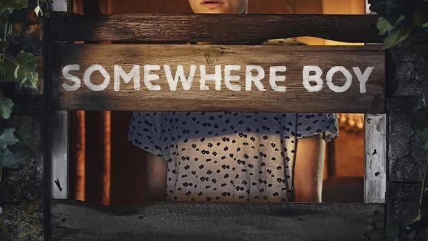 Ein junger Mann blickt besorgt durch eine hölzerne Fensteröffnung mit dem Schriftzug „Somewhere Boy“.