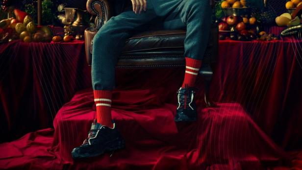 Ein Mann sitzt in einem Sessel, umgeben von Kerzenleuchtern und einem Tisch voller Früchte.