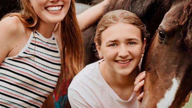 Zwei junge Frauen umarmen ein braunes Pferd; Filmtitel „Mein Pferdesommer“.