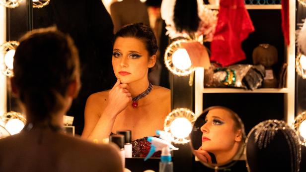 Eine Frau mit starkem Make-up blickt in einem beleuchteten Spiegel nachdenklich in die Ferne.