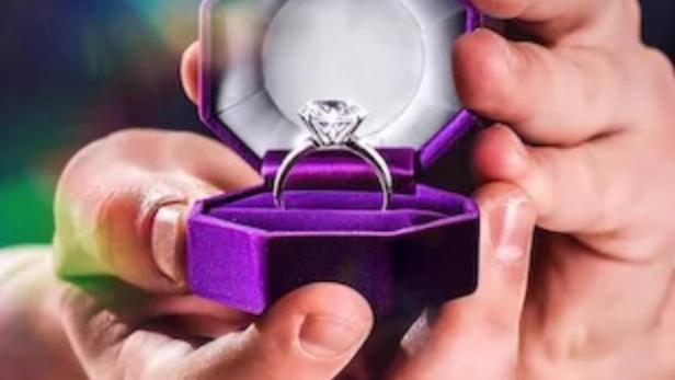 Ein Mann hält eine geöffnete, lilafarbene Schmuckschatulle mit einem Diamantring für „Love is Blind UK“.