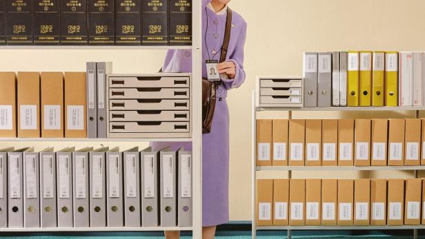 Eine junge Frau in einem lila Kostüm steht vor einem Regal voller Akten und Gesetzbücher.