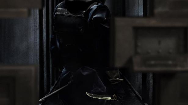 Ein Mann mit einer Narbe im Gesicht, der einen Dolch hält, im Hintergrund das Logo von „House of Ninjas“.