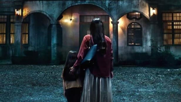 Eine Frau und ein Kind stehen vor einem unheimlichen Haus, in einem Fenster ist eine geisterhafte Gestalt zu sehen.