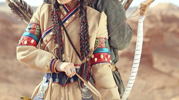 Eine junge Frau und ein Mann, beide in traditioneller mongolischer Kleidung, mit Pfeil und Bogen in der Wüste.