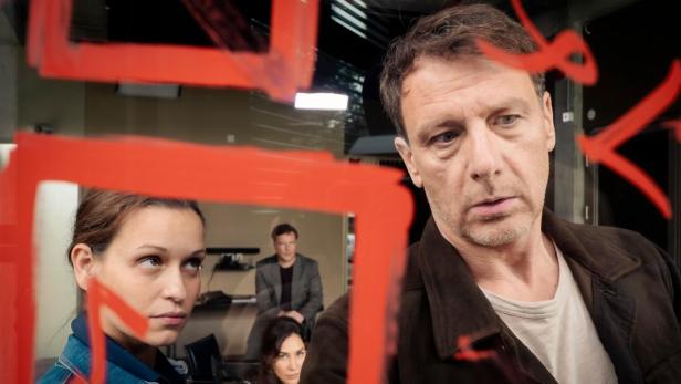 Szene aus dem deutschen Fernsehfilm „Dünentod – Ein falsches Spiel“ mit einem Mann und einer Frau vor einer beschmierten Scheibe.