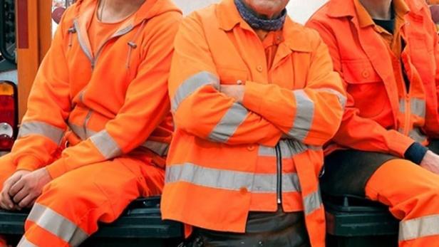 Drei Männer in orangefarbenen Overalls sitzen auf einer Mülltonne, Plakat für „Die Drei von der Müllabfuhr: Baby an Bord“.