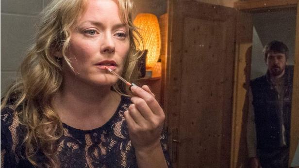 Eine Frau trägt Lippenstift vor einem Spiegel auf, im Hintergrund ein Mann.