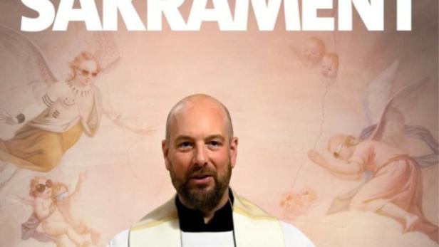 Ein Priester steht vor einem Wandgemälde mit Engeln und dem Schriftzug „Himmel Herrgott Sakrament“.