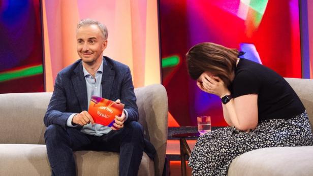 Ein Mann hält eine Karte mit der Aufschrift „Lass dich überwachen“, während eine Frau daneben ihr Gesicht verdeckt.