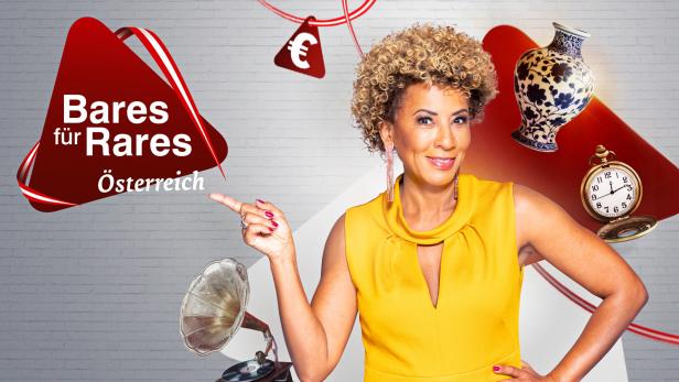 Arabella Kiesbauer präsentiert „Bares für Rares Österreich“ vor einem grauen Hintergrund mit Antiquitäten.