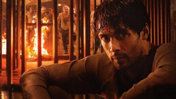 Ein Mann sitzt vor einem Gitter in „Celda 211“, während im Hintergrund ein Feuer brennt.