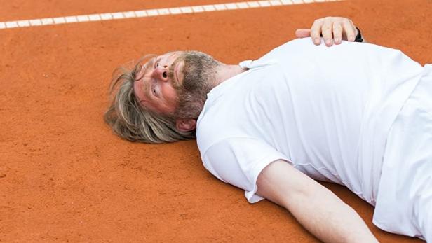 Ein Mann liegt erschöpft mit einem Tennisschläger auf einem Tennisplatz, Filmposter „Los Paul“.