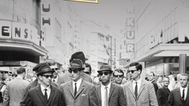 Männer in Anzügen und Hüten gehen in Köln die Straße entlang.