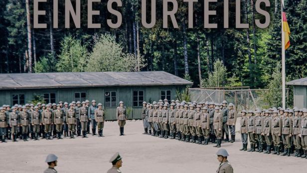 Soldaten in Uniform stehen auf einem Exerzierplatz vor Baracken und einer Deutschlandflagge.