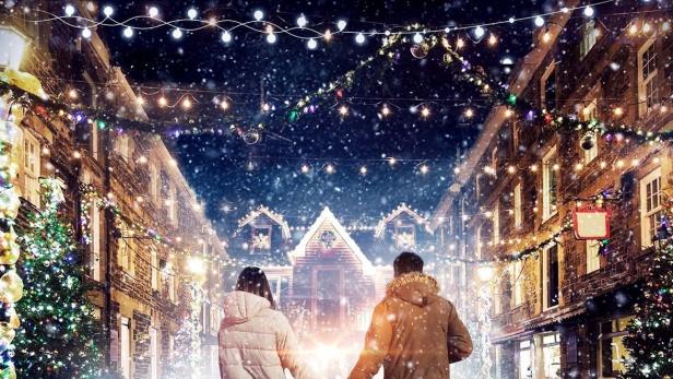 Ein Paar geht Händchen haltend durch eine verschneite Gasse, geschmückt mit Lichtern, vor dem „Holly Day Inn“.